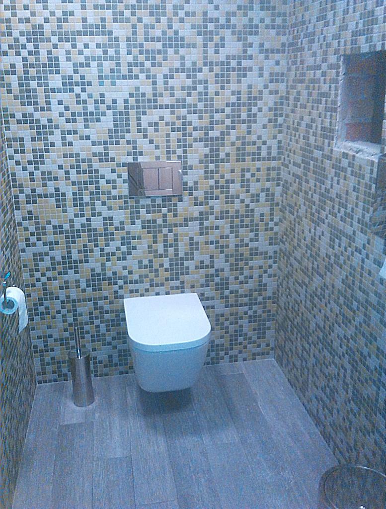 Wykończona łazienka z mozaiką w odcieniach szarości, beżu i bieli. Podwieszana toaleta, drewnopodobna podłoga, metalowe akcesoria łazienkowe.