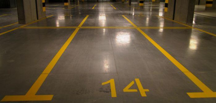 Szara powierzchnia parkingu podziemnego z żółtymi liniami wyznaczającymi miejsca postojowe i numerem '14' na jednym ze stanowisk.
