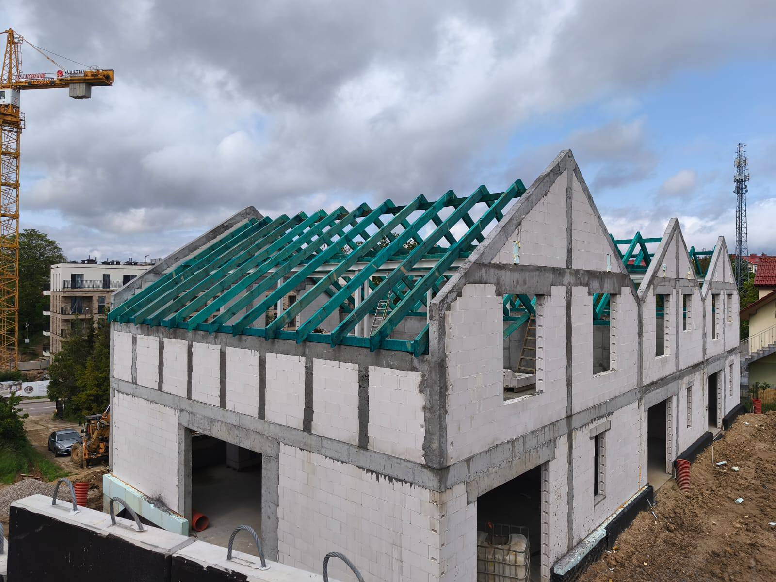 Wznoszenie domu z białych bloczków: ściany parteru i poddasza, zielona konstrukcja dachu. Na drugim planie żuraw budowlany i wieża telekomunikacyjna.