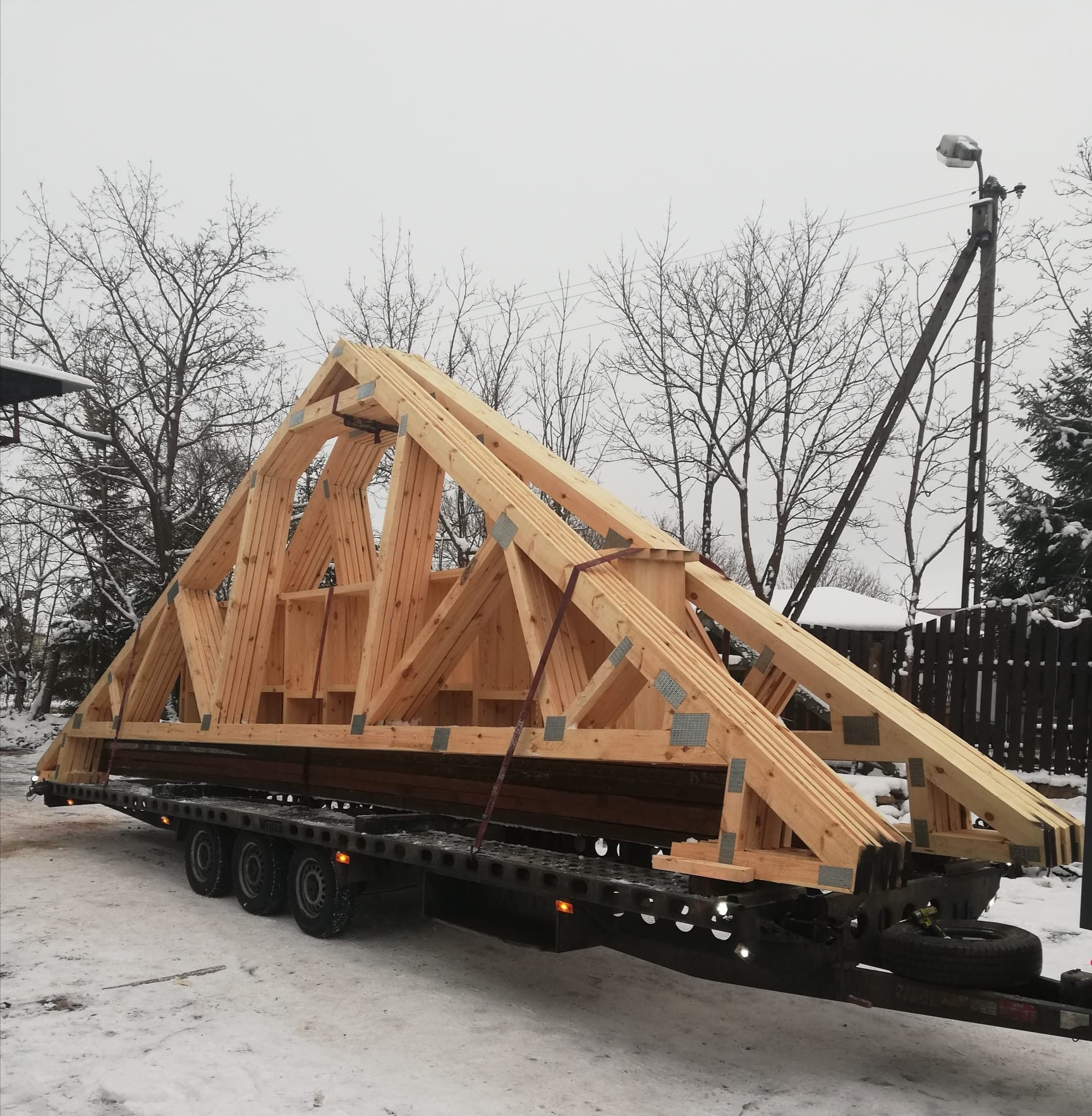 Gotowa więźba dachowa z jasnego drewna, zabezpieczona pasami, transportowana na przyczepie w zimowej scenerii. Widoczne metalowe łączniki i elementy konstrukcyjne.