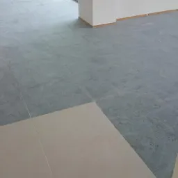 Szara, kwadratowa płytka podłogowa ułożona w dużym pomieszczeniu, częściowo pokryta jasną płytką. Widoczny fragment kolumny i ściany.