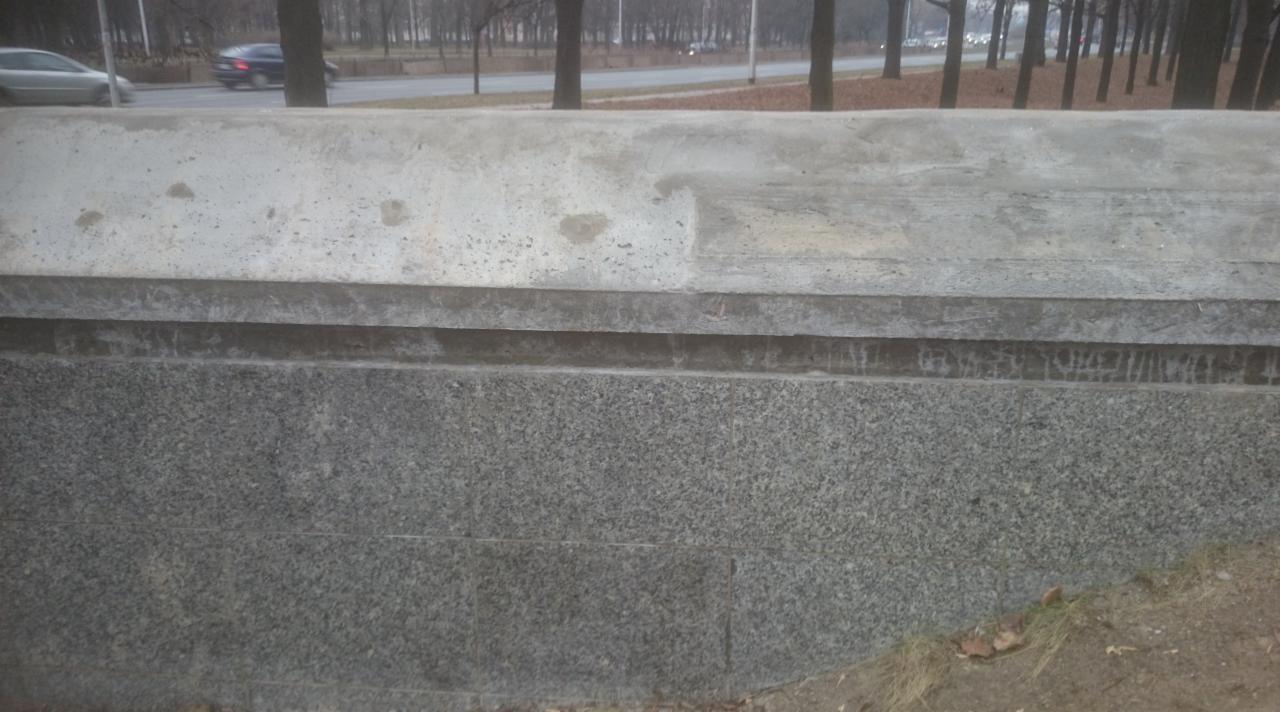 Szara, granitowa elewacja zewnętrzna z betonowym wykończeniem, widoczna z bliska. Tło stanowią drzewa i ulica z przejeżdżającymi samochodami.