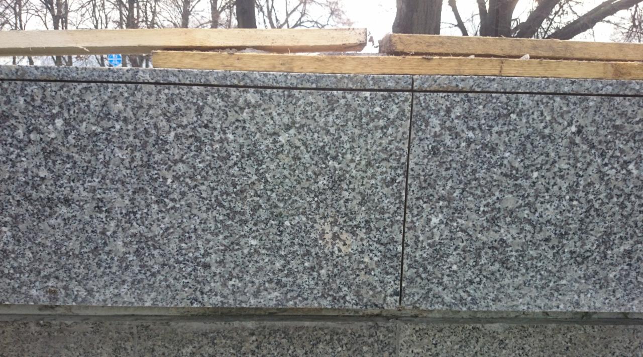 Szara, granitowa okładzina ściany zewnętrznej z drewnianymi elementami montażowymi u góry, widoczne drzewa w tle.