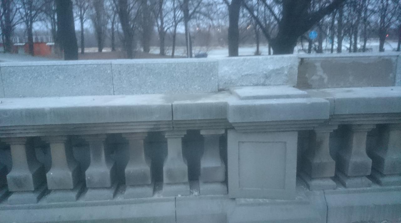 Kamienna balustrada z filarami na tle drzew, utrzymana w szarościach. Widoczne elementy konstrukcyjne i detale architektoniczne.