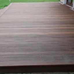 Nowoczesny taras z brązowych desek kompozytowych, widok z góry, z fragmentem trawnika i elewacji budynku w tle. Deski ułożone równolegle.