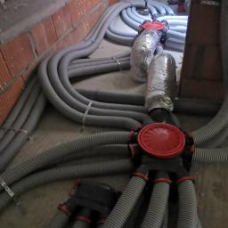 C.L.Klima - Szare, karbowane rury wentylacyjne z czerwonymi łącznikami rozchodzą się po podłodze, część rur owinięta folią aluminiową, widoczna ściana z cegły i ocieplenie pianką.