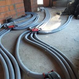 C.L.Klima - Szare, karbowane rury wentylacyjne rozchodzą się promieniście od czerwonych rozdzielaczy na surowym betonowym podłożu w trakcie montażu w budynku z cegły.