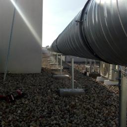 C.L.Klima - Długi, srebrny kanał wentylacyjny na dachu pokrytym żwirem, wsparty na metalowych wspornikach, z jednostkami zewnętrznymi klimatyzacji w tle i narzędziami do montażu widocznymi w lewym dolnym rogu.