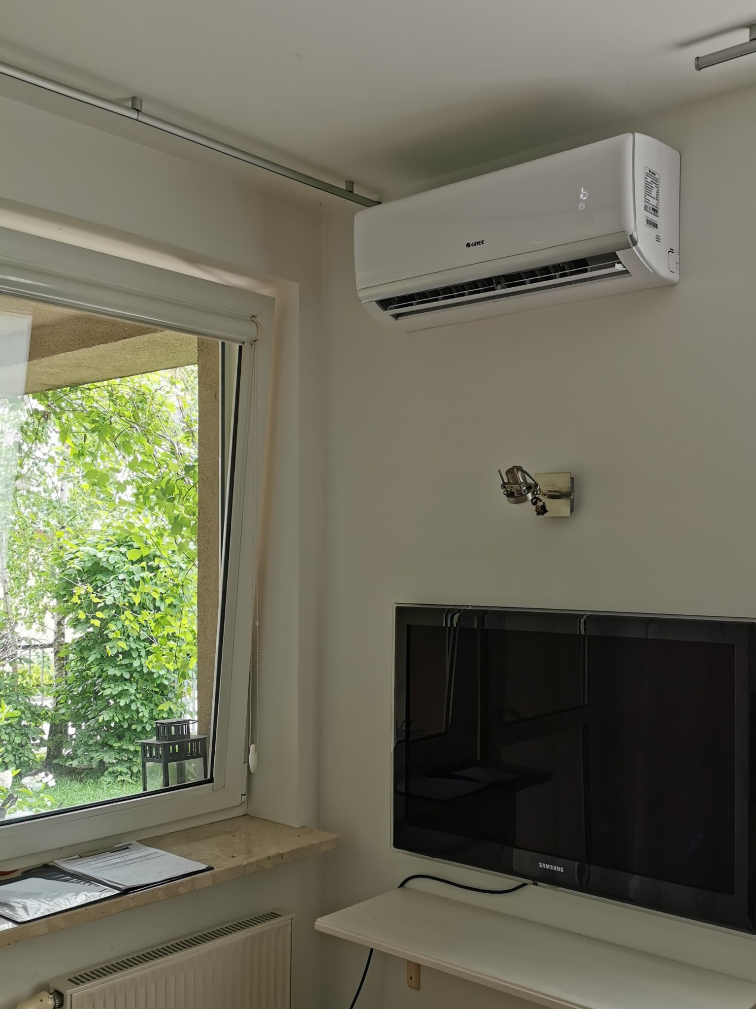 Biały klimatyzator marki Gree zamontowany na ścianie w pokoju z oknem i telewizorem Samsung.