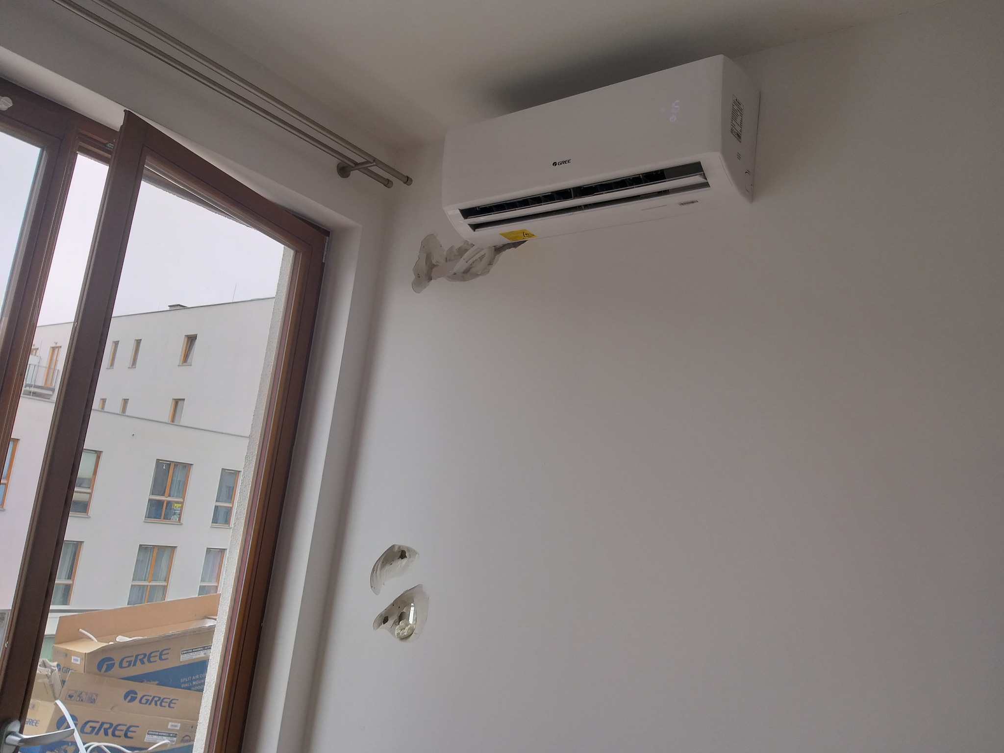Biały klimatyzator Gree zamontowany na ścianie, widoczne ślady po instalacji i kartony klimatyzatorów Gree w tle za oknem.