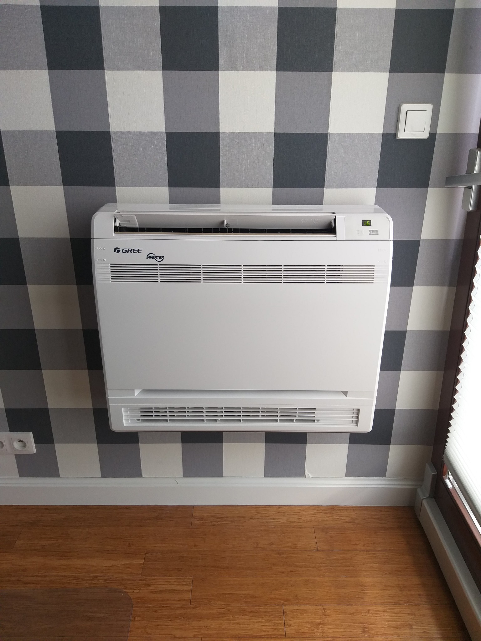 Biała, przypodłogowa jednostka klimatyzacyjna Gree Inverter zamontowana na ścianie w kratę, z widocznym wyświetlaczem temperatury i drewnianą podłogą.