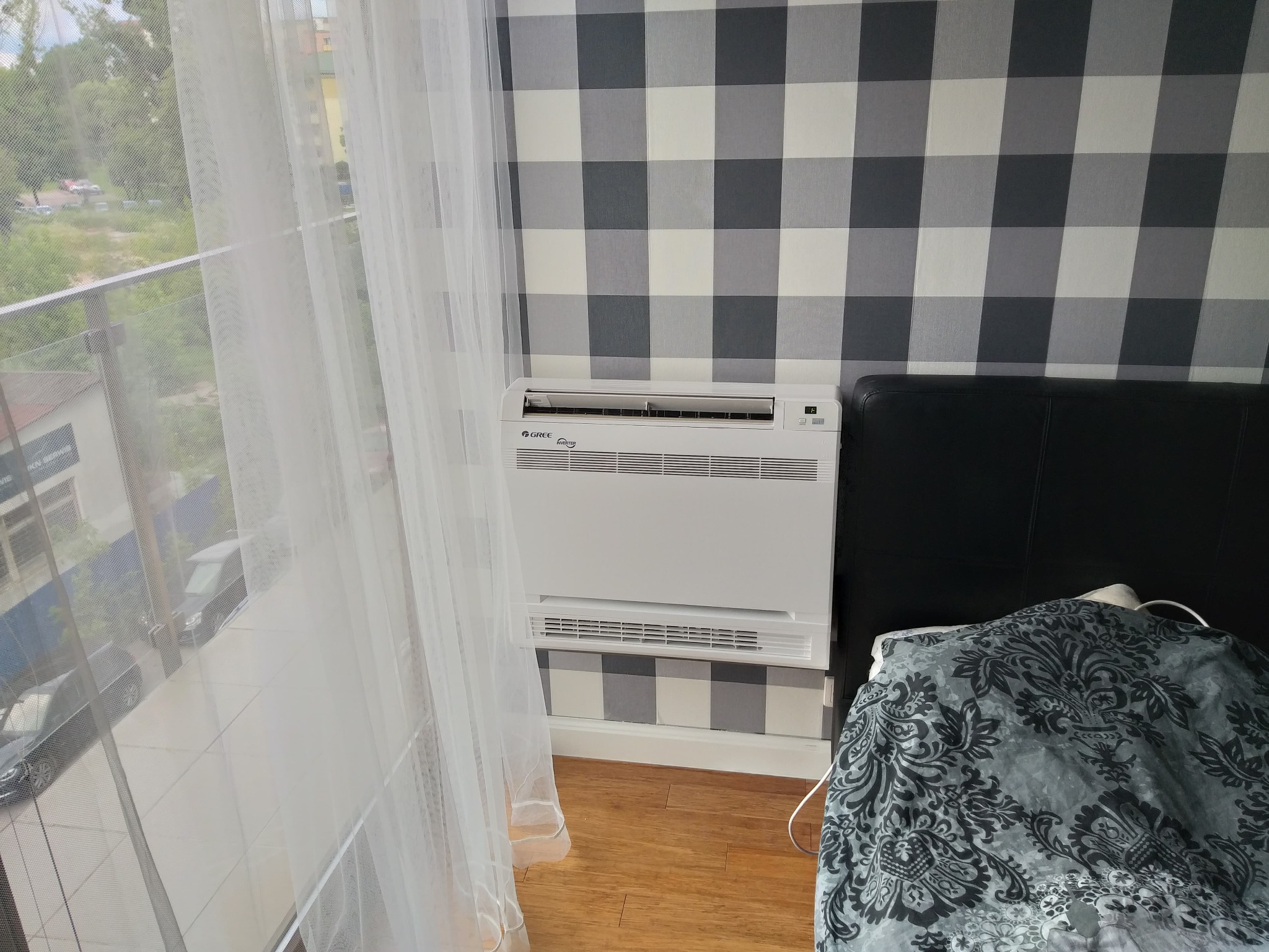 Biała, przypodłogowa jednostka klimatyzacyjna Gree z widocznym logo, zamontowana w pokoju z tapetą w szaro-białą kratę, obok łóżka z ciemnym zagłówkiem i wzorzystą pościelą, z widokiem na zewnątrz...