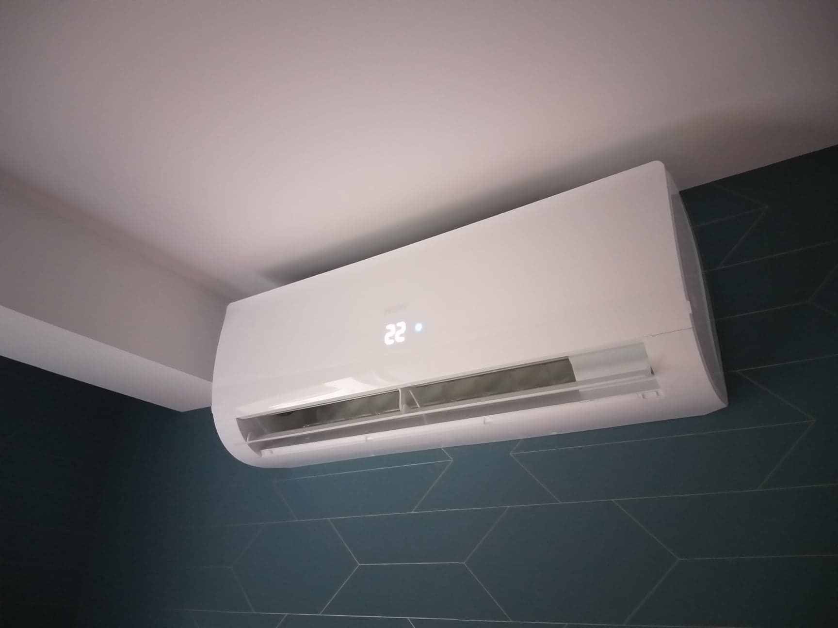Biała jednostka klimatyzacji Haier zamontowana na ścianie z ciemnymi, geometrycznymi płytkami. Na wyświetlaczu widoczna temperatura 22 stopnie.