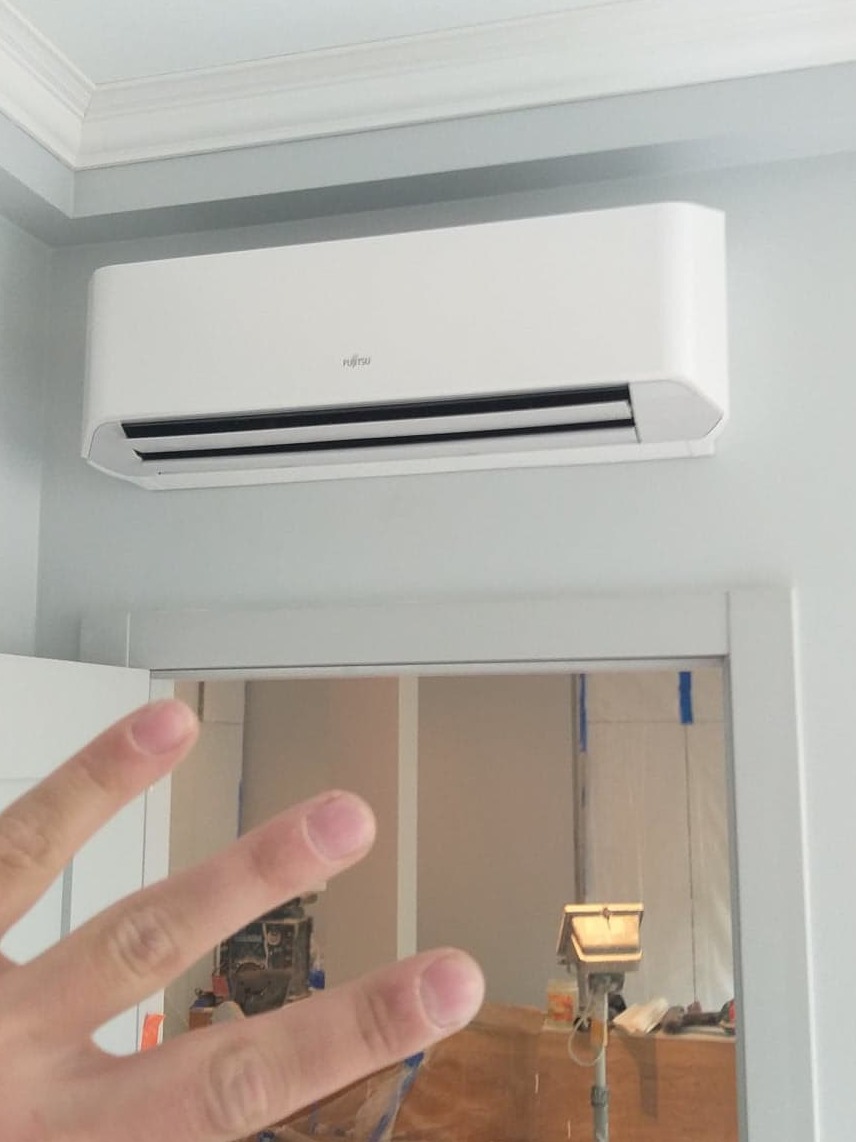 Biała jednostka wewnętrzna klimatyzacji Fujitsu zamontowana na ścianie w jasnym pomieszczeniu z widocznym fragmentem dłoni w kadrze i wnętrzem w trakcie remontu w tle.