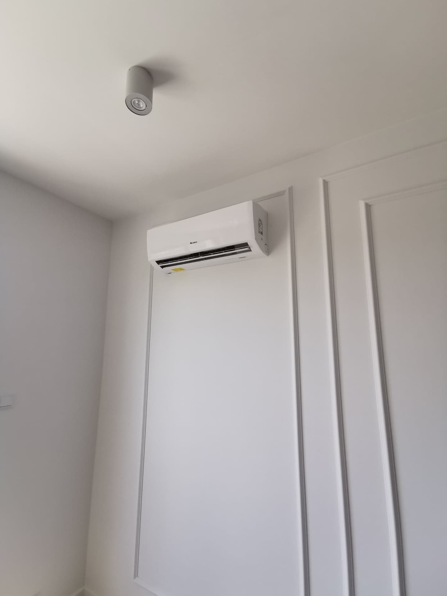 Biały klimatyzator ścienny marki Gree zamontowany w rogu pokoju z dekoracyjnymi panelami ściennymi i punktowym oświetleniem sufitowym.