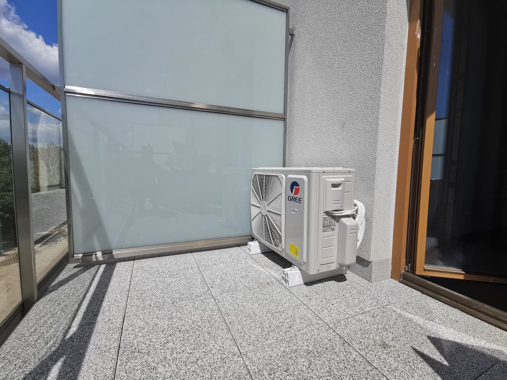Biała jednostka zewnętrzna klimatyzacji Gree zamontowana na balkonie z szarą, granitową posadzką, widok z boku, częściowo widoczne przeszklone balustrady.
