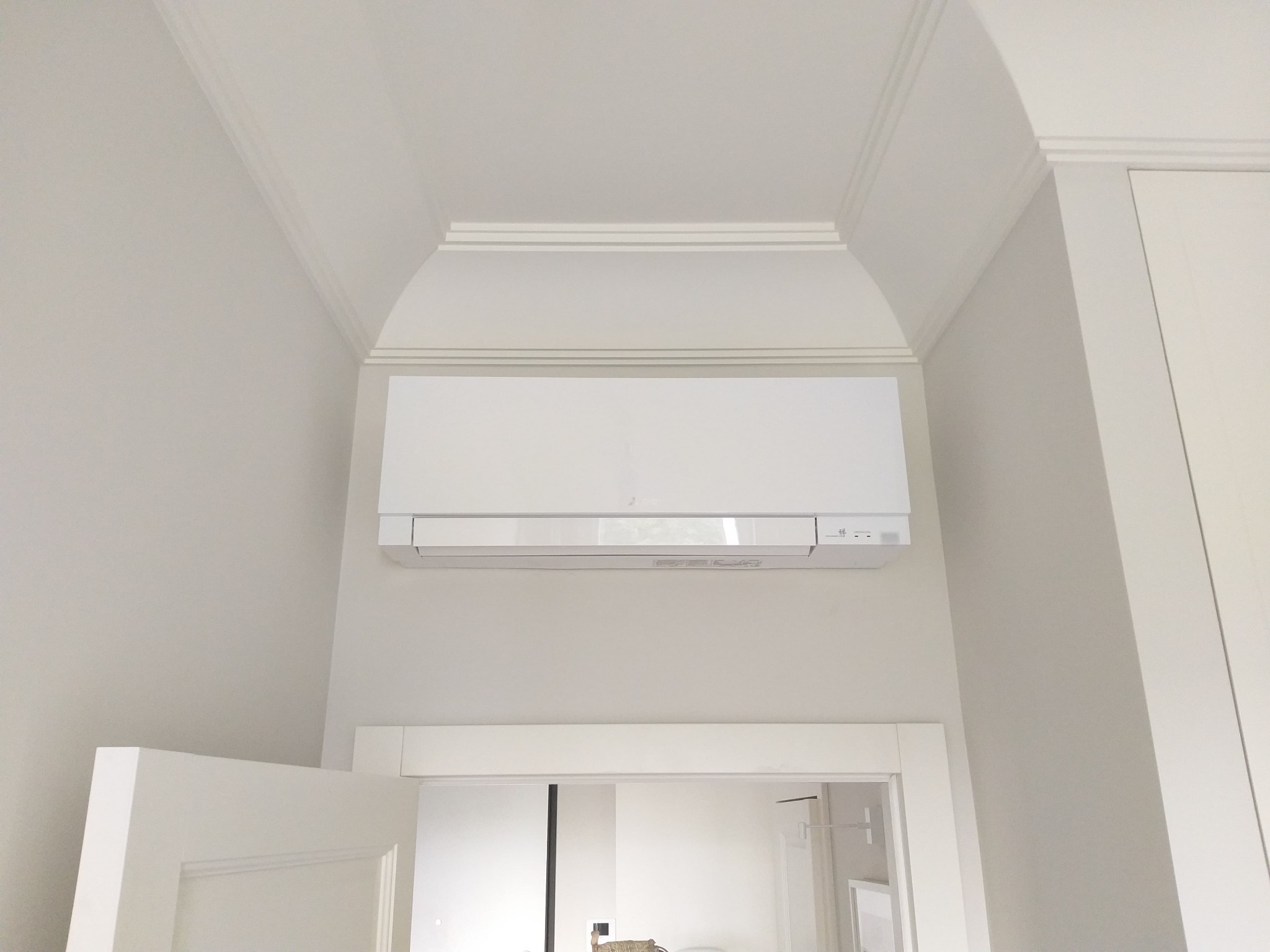 Biała klimatyzacja Mitsubishi Electric zamontowana nad drzwiami w jasnym wnętrzu z dekoracyjną listwą sufitową.