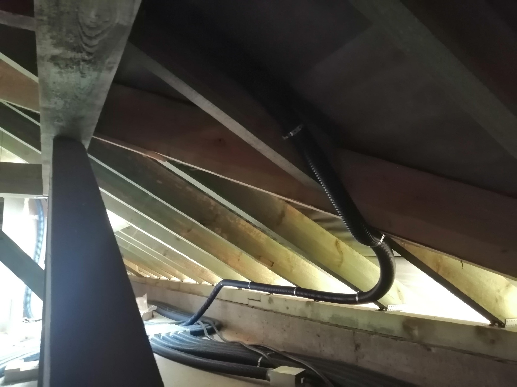 Czarna, karbowana rura wentylacyjna zamontowana pod skośnym dachem z drewnianych belek, zabezpieczona metalowymi opaskami zaciskowymi.