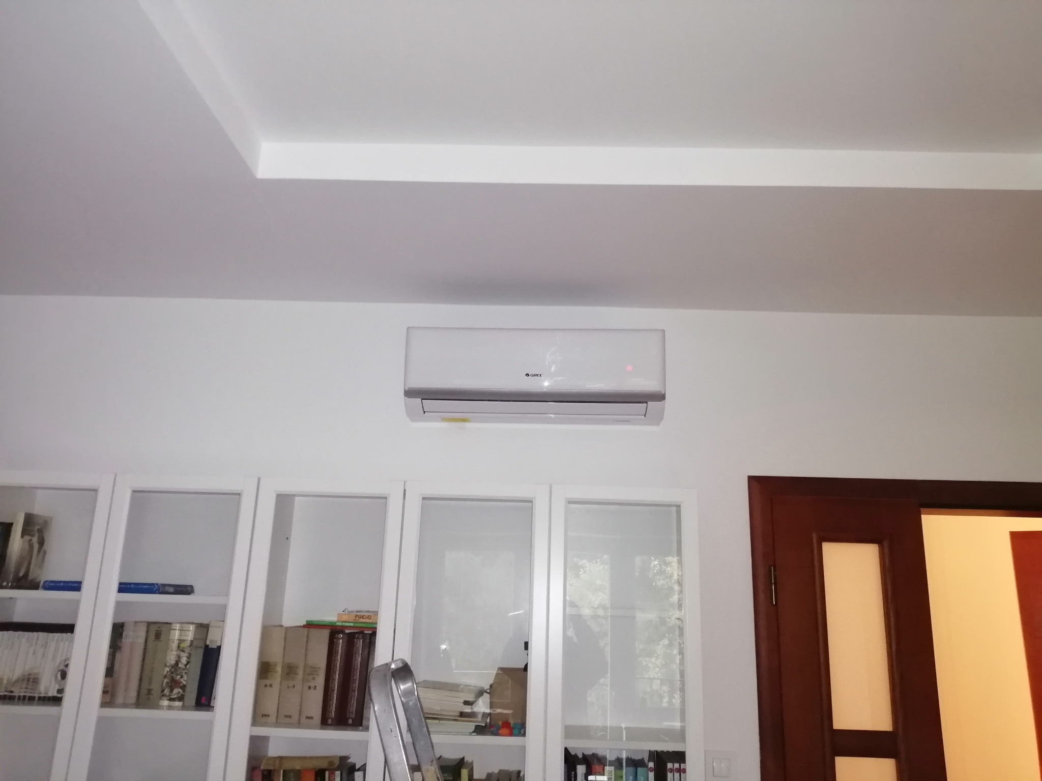 Biały klimatyzator marki Gree zamontowany na ścianie w pokoju w Warszawie, widoczny regał z książkami i fragment drzwi.