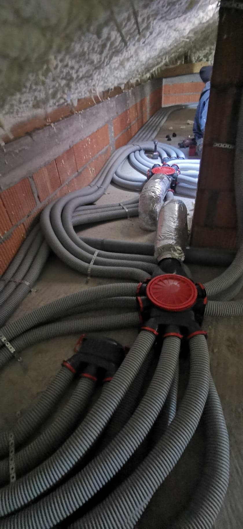 Szare, karbowane rury wentylacyjne z czerwonymi łącznikami rozchodzą się po podłodze, część rur owinięta folią aluminiową, widoczna ściana z cegły i ocieplenie pianką.