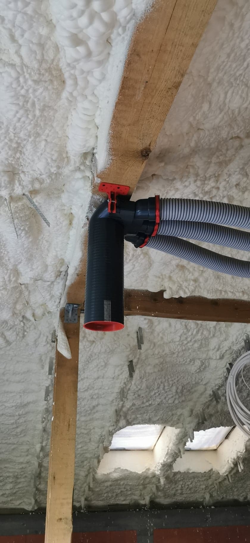 Szare, karbowane przewody wentylacyjne podłączone do czarnego pionowego kanału z pomarańczowym wnętrzem, zamontowane pod drewnianą belką stropową i ocieplonym pianą dachem.