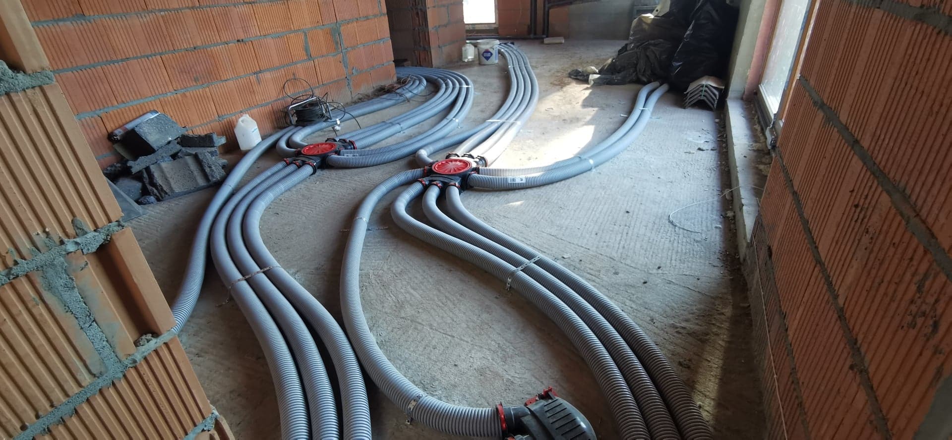 Szare, karbowane rury wentylacyjne rozchodzą się promieniście od czerwonych rozdzielaczy na surowym betonowym podłożu w trakcie montażu w budynku z cegły.
