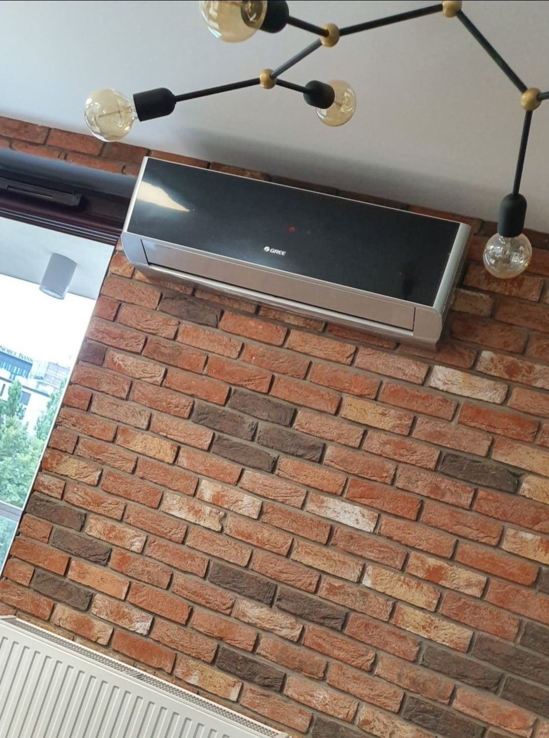 Czarny klimatyzator marki Gree zamontowany na ścianie z cegły, oświetlony nowoczesną lampą z żarówkami Edisona.