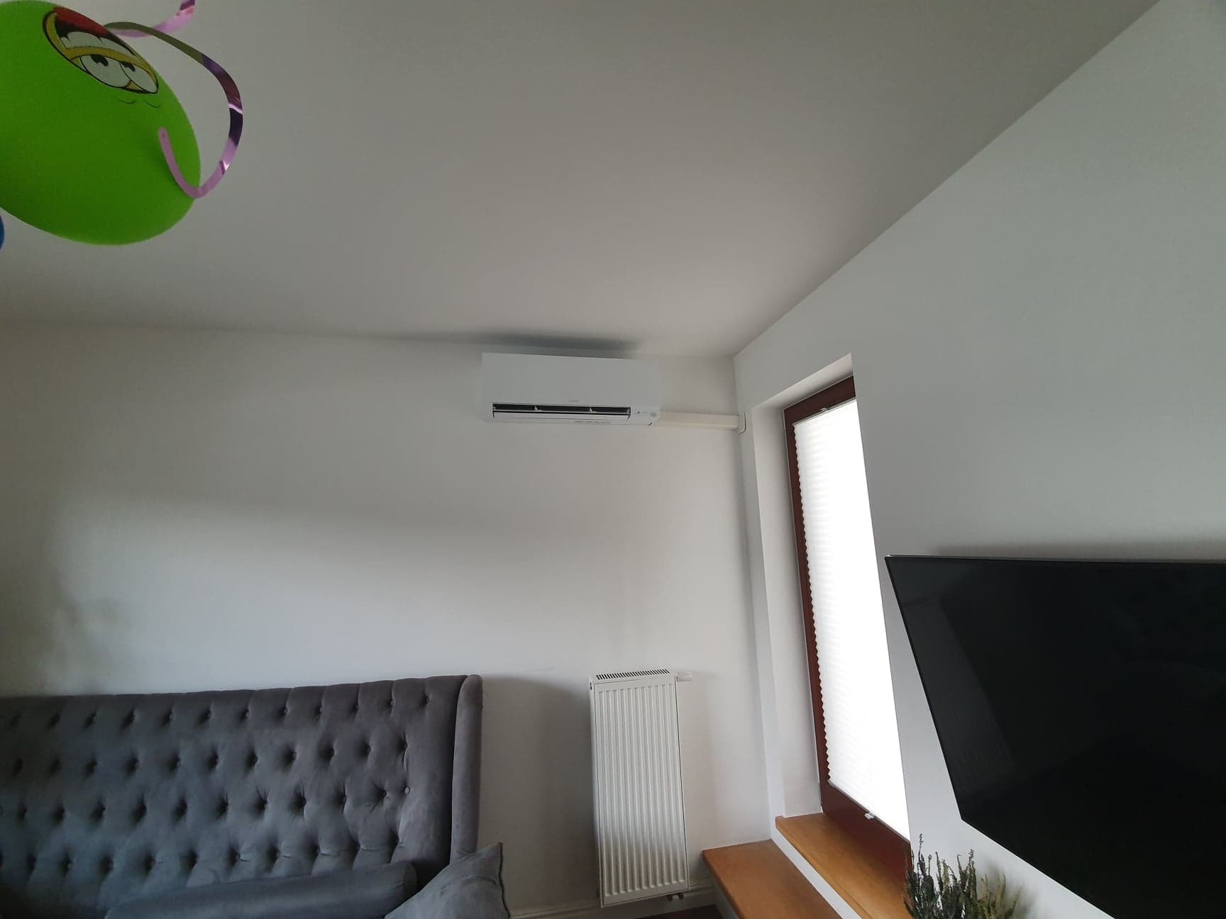 Biały klimatyzator ścienny zamontowany w pokoju z szarą sofą, białym grzejnikiem i oknem z żaluzjami, z zielonym balonem z twarzą w lewym górnym rogu.