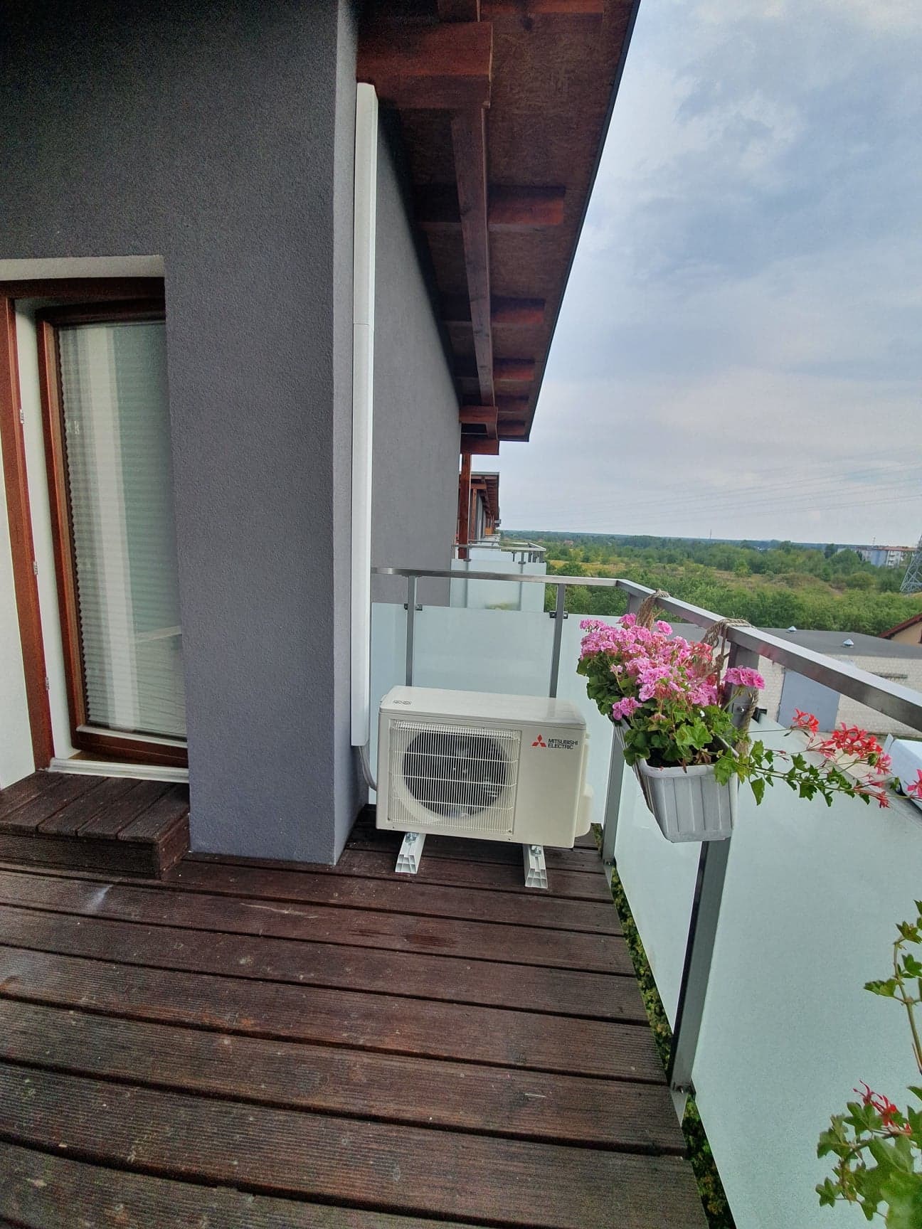 Jednostka zewnętrzna klimatyzatora Mitsubishi Electric zamontowana na balkonie z drewnianą podłogą, obok doniczki z różowymi kwiatami, z widokiem na zielony krajobraz.