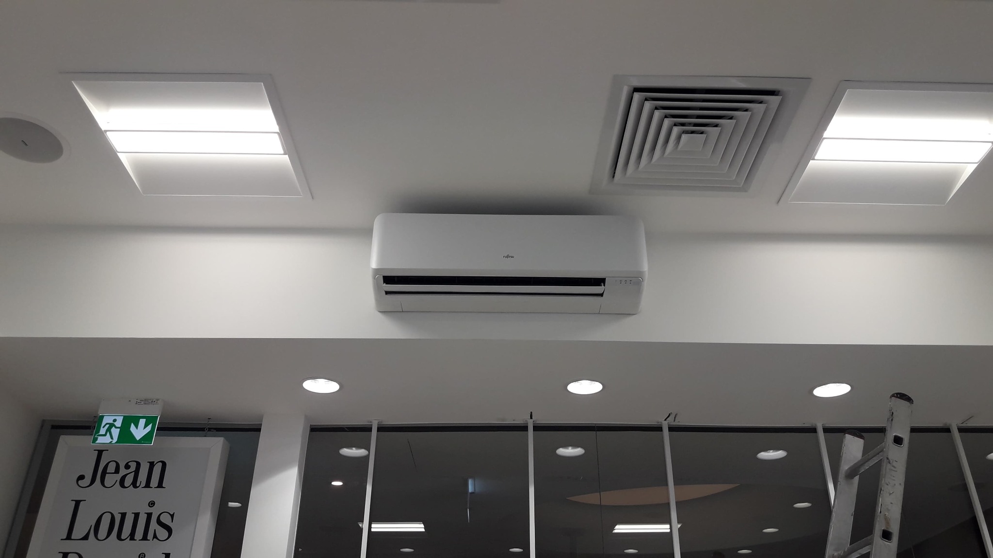 Biała jednostka wewnętrzna klimatyzatora Fujitsu zamontowana na suficie w pomieszczeniu biurowym, widoczne oświetlenie sufitowe i fragment drabiny.