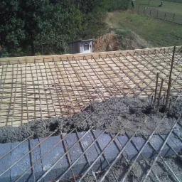 Zbrojenie fundamentu budynku z częściowo wylanym betonem, widoczne pionowe pręty i elementy drewnianego szalunku, w tle zielony teren z ogrodzeniem.