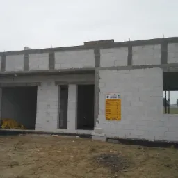 Surowy stan budynku jednorodzinnego z betonu komórkowego, widoczne otwory okienne i drzwiowe, na ścianie wisi żółta tablica informacyjna, w tle widoczne drzewa.