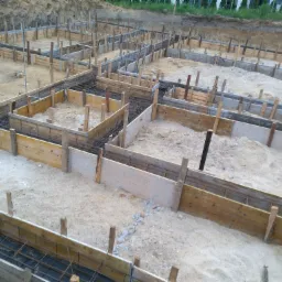 Przygotowanie szalunku pod fundamenty z widocznym zbrojeniem, teren budowy z ziemią i drewnianymi elementami konstrukcyjnymi.