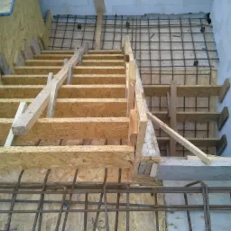 Konstrukcja szalunku schodów z płyty OSB, zbrojenie stalowe widoczne w tle i u dołu, surowy beton na ścianie.