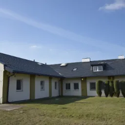 Budynek z ciemnym dachem z dachówki, widoczne okna dachowe, rynny i pionowe tuje wzdłuż ściany.