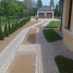 Granitowa kostka tworząca ścieżkę i podjazd do garażu, otoczone trawnikiem i tujami, widok z perspektywy domu.