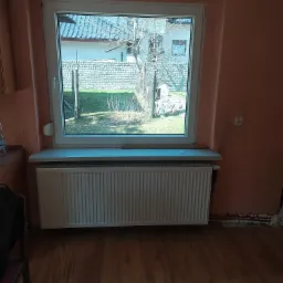 Biały grzejnik panelowy zamontowany pod oknem w pokoju z pomarańczowymi ścianami i drewnianą podłogą; widoczne fragmenty instalacji grzewczej w ścianie.