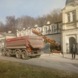 Ciężarówka wywrotka z podniesioną skrzynią na tle budynku w trakcie prac, widoczny mężczyzna w kurtce i czapce oraz gruz i elementy konstrukcyjne.