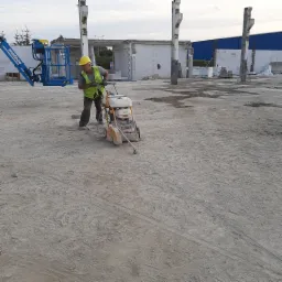 Pracownik w żółtym kasku przecina betonową nawierzchnię za pomocą przecinarki CEDIMA, w tle częściowo rozebrany budynek z białego bloczka i podnośnik koszowy.