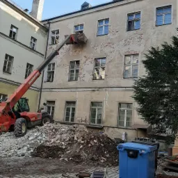 Czerwona ładowarka teleskopowa MANISCOPIC podnosi gruz z wyburzonej ściany na tle budynku z odsłoniętą cegłą i niebieskim kontenerem na odpady.