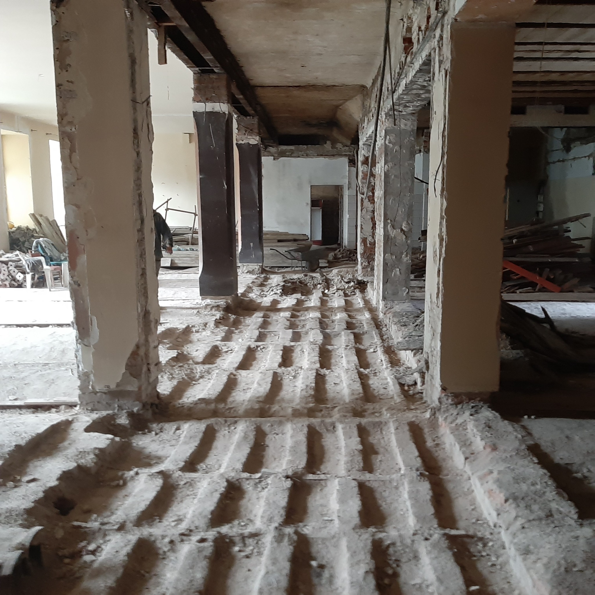 Wnętrze budynku w trakcie demontażu, widoczne fragmenty ścian, odsłonięta konstrukcja podłogi z pozostałościami materiałów budowlanych, w tle taczka i składowane elementy.