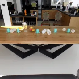 Stół jadalny z blatem z jasnego drewna i geometryczną, czarną podstawą, udekorowany turkusowymi świecami, białymi kulami dekoracyjnymi, dzbankiem z wodą i misą z owocami, na tle nowoczesnej kuchni.