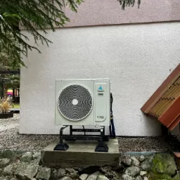 Zewnętrzna jednostka pompy ciepła Vesser Alterius DC Inverter, zamontowana na betonowym fundamencie przed budynkiem o jasnej elewacji z elementami zieleni.