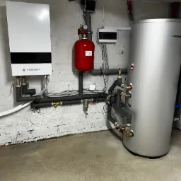 Biały, wiszący piec grzewczy Vesser z podłączonymi rurami, czerwony zbiornik wyrównawczy i srebrny bojler w kotłowni z surowymi, pomalowanymi na biało ścianami z cegły.