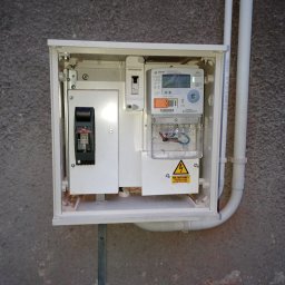 MEDIA-INSTAL SPÓŁKA Z OGRANICZONĄ ODPOWIEDZIALNOŚCIĄ - Zamknięta skrzynka elektryczna z licznikiem i bezpiecznikami na szarej ścianie. Widoczne białe rury instalacyjne. Tabliczka ostrzegawcza o urządzeniu elektrycznym.