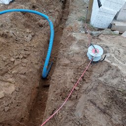 MEDIA-INSTAL SPÓŁKA Z OGRANICZONĄ ODPOWIEDZIALNOŚCIĄ - Wykop w ziemi z niebieską rurą karbowaną i szpulą z kablem. Przygotowanie do instalacji elektrycznej w gruncie.