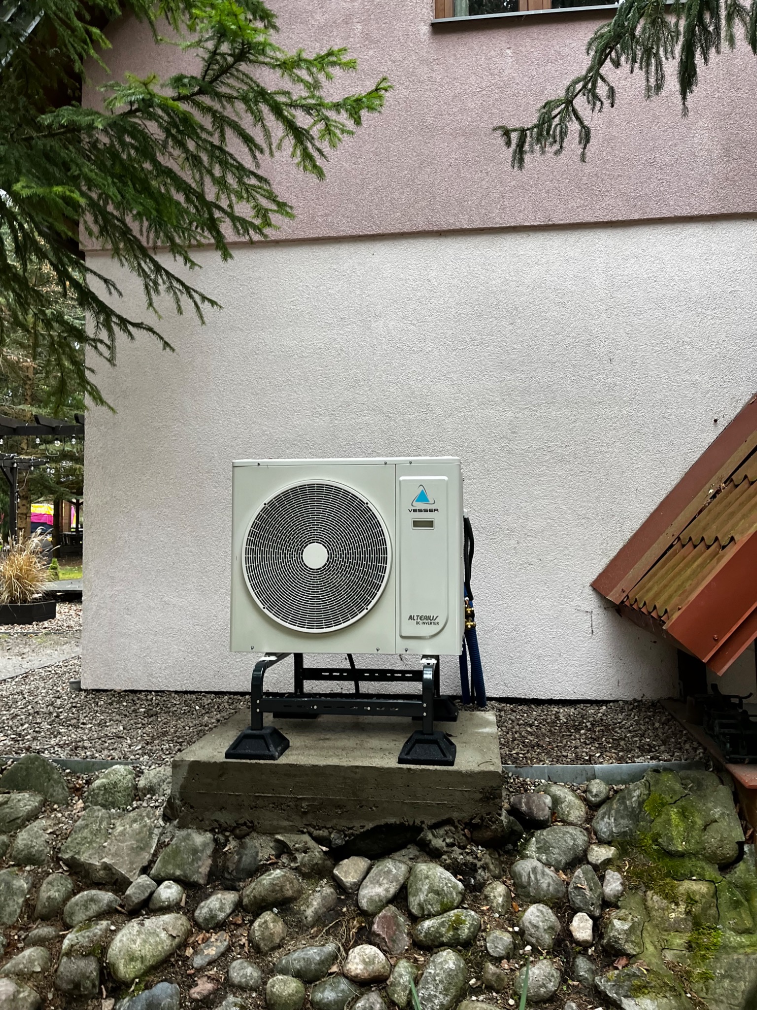 Zewnętrzna jednostka pompy ciepła Vesser Alterius DC Inverter, zamontowana na betonowym fundamencie przed budynkiem o jasnej elewacji z elementami zieleni.