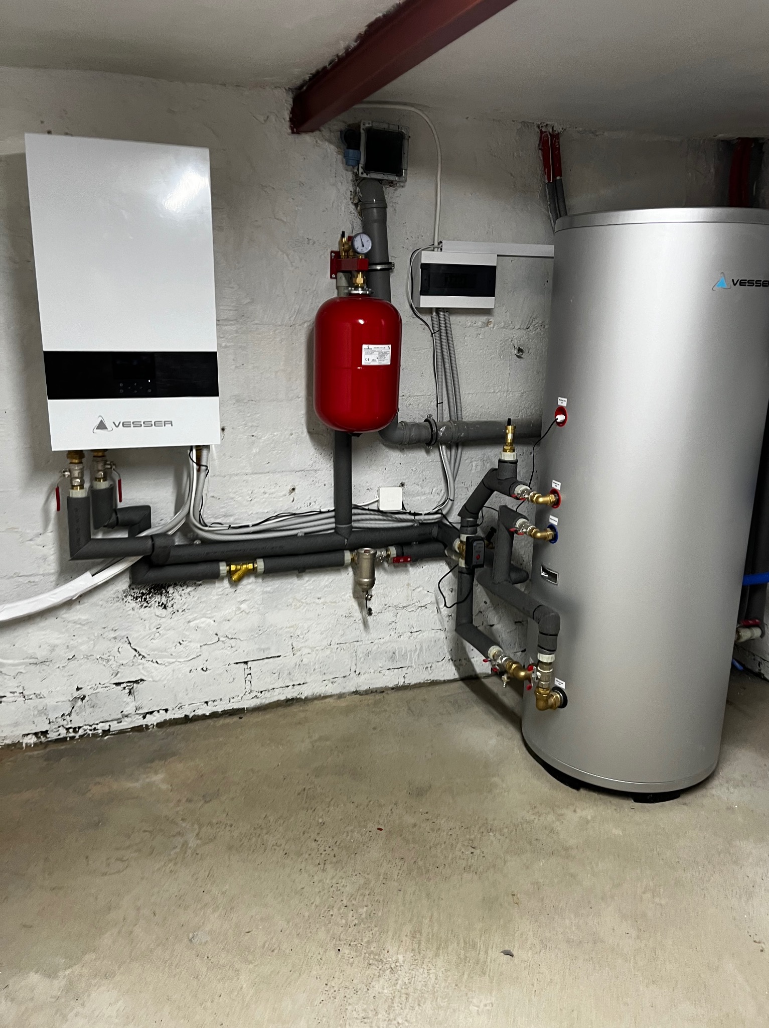 Biały, wiszący piec grzewczy Vesser z podłączonymi rurami, czerwony zbiornik wyrównawczy i srebrny bojler w kotłowni z surowymi, pomalowanymi na biało ścianami z cegły.