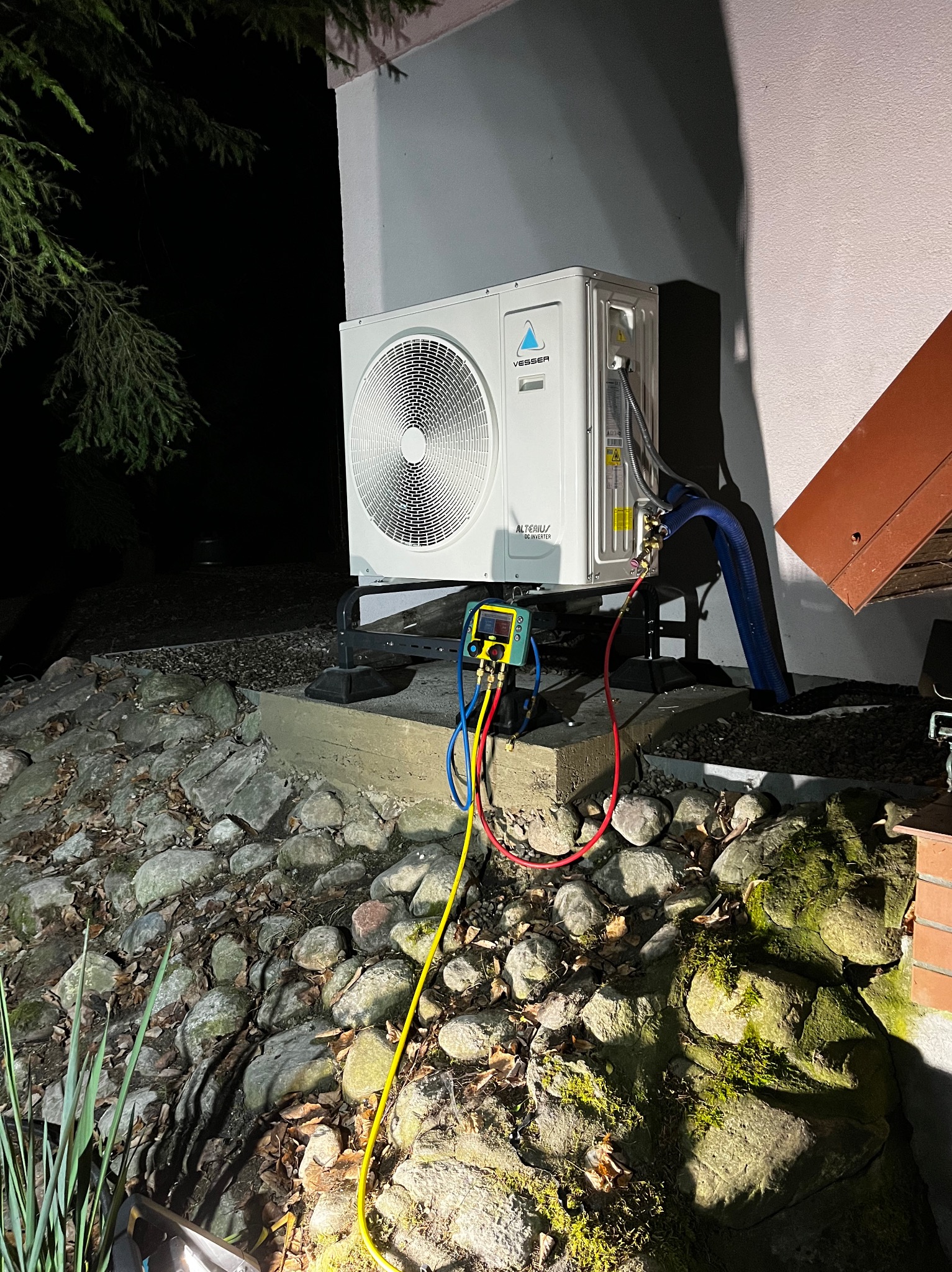 Biała jednostka zewnętrzna klimatyzacji marki Vesser, podłączona do manometrów z kolorowymi przewodami, stojąca na betonowym fundamencie w ogrodzie z kamieniami.