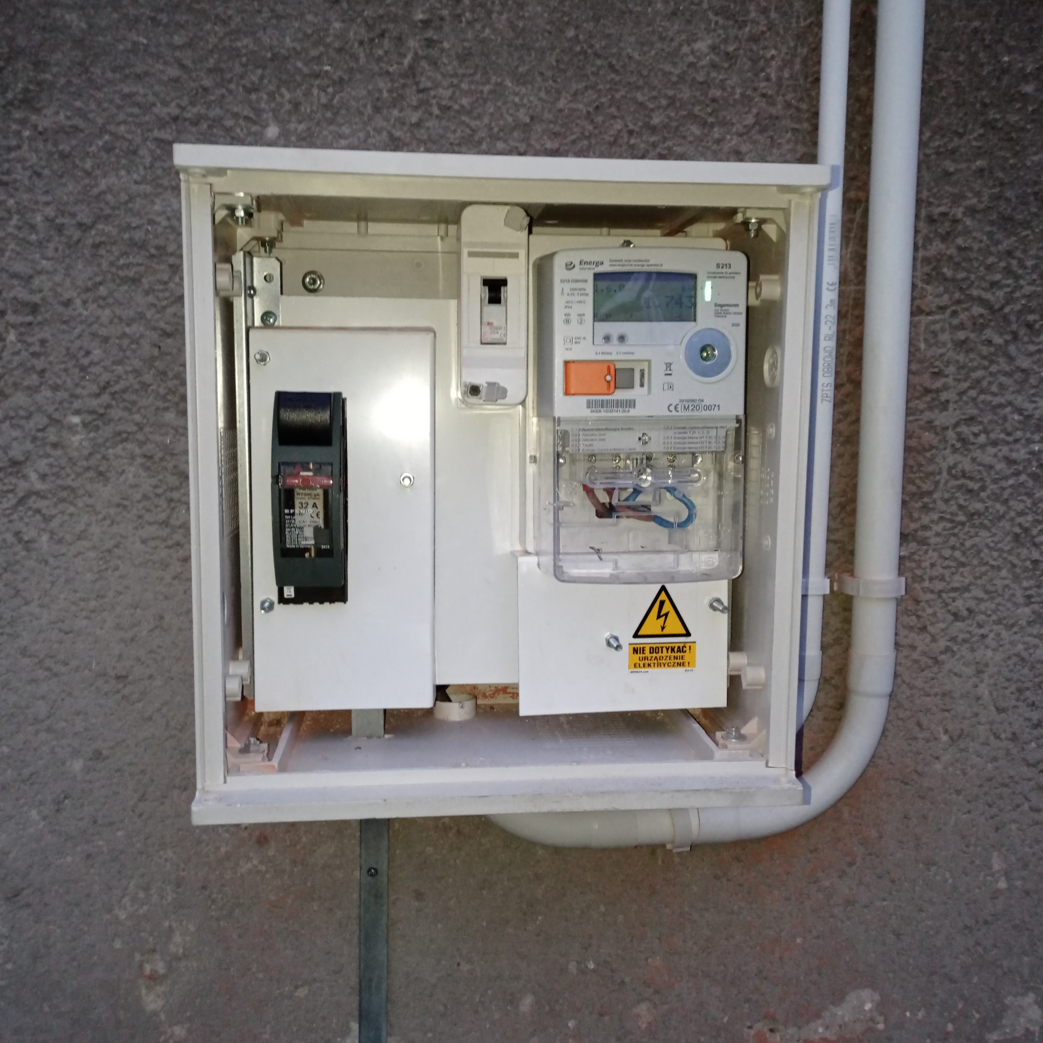 Zamknięta skrzynka elektryczna z licznikiem i bezpiecznikami na szarej ścianie. Widoczne białe rury instalacyjne. Tabliczka ostrzegawcza o urządzeniu elektrycznym.